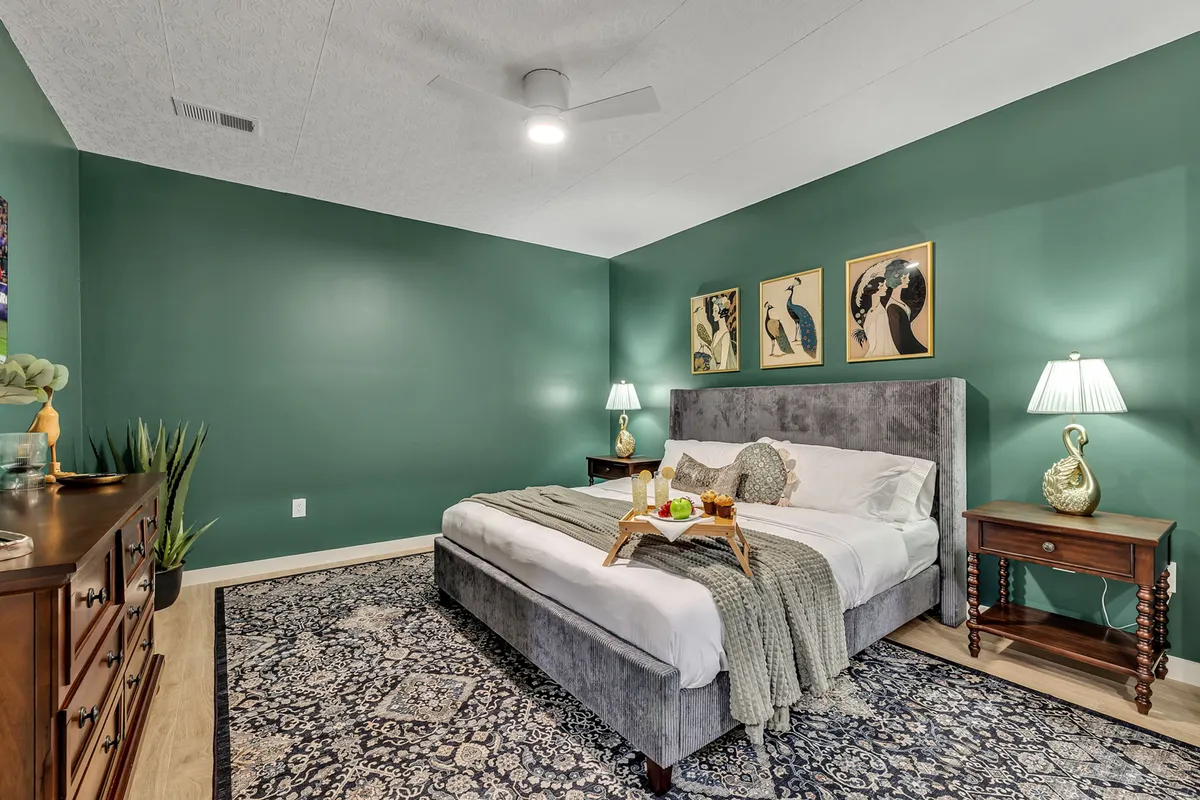 A tranquil bedroom scene with a bed dressed in decorative pillows, soft bedding, and a bedside table with a lamp and a decorative plant. Artwork hangs on the wall above the bed, contributing to the room's calming aesthetic.