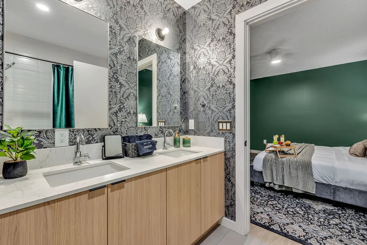 A sophisticated bathroom scene with a dual sink vanity, large mirrors, elegant floral wallpaper, a green accent wall, and a partial view into the adjoining bedroom. The lighting is soft and calming, evoking a spa-like ambiance.