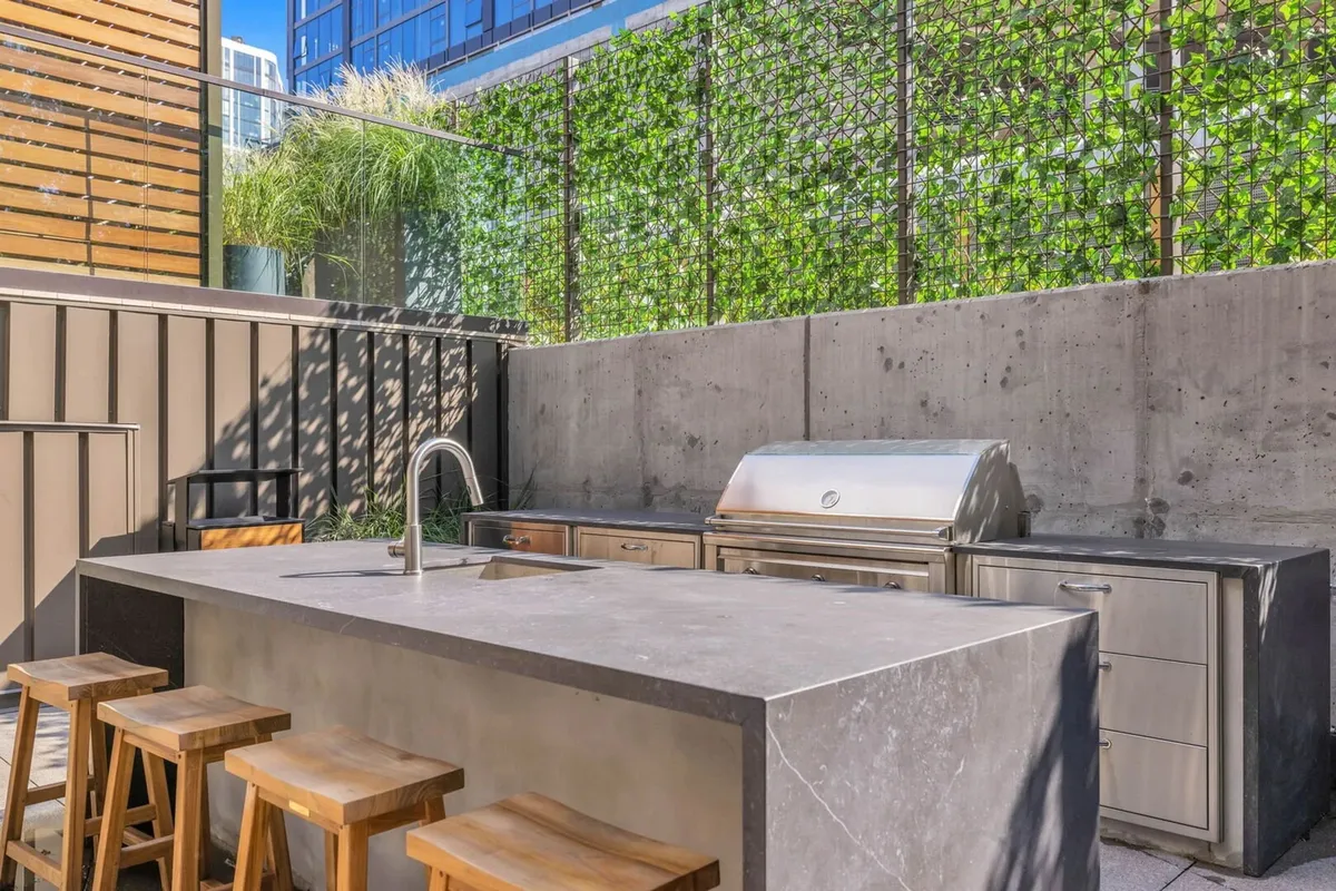 A stylish outdoor kitchen setup with a concrete countertop, bar stools, and surrounding greenery, inviting guests to gather for barbecues or sunset dinners. This space is designed for group meals, celebration, and socializing in an open-air environment.