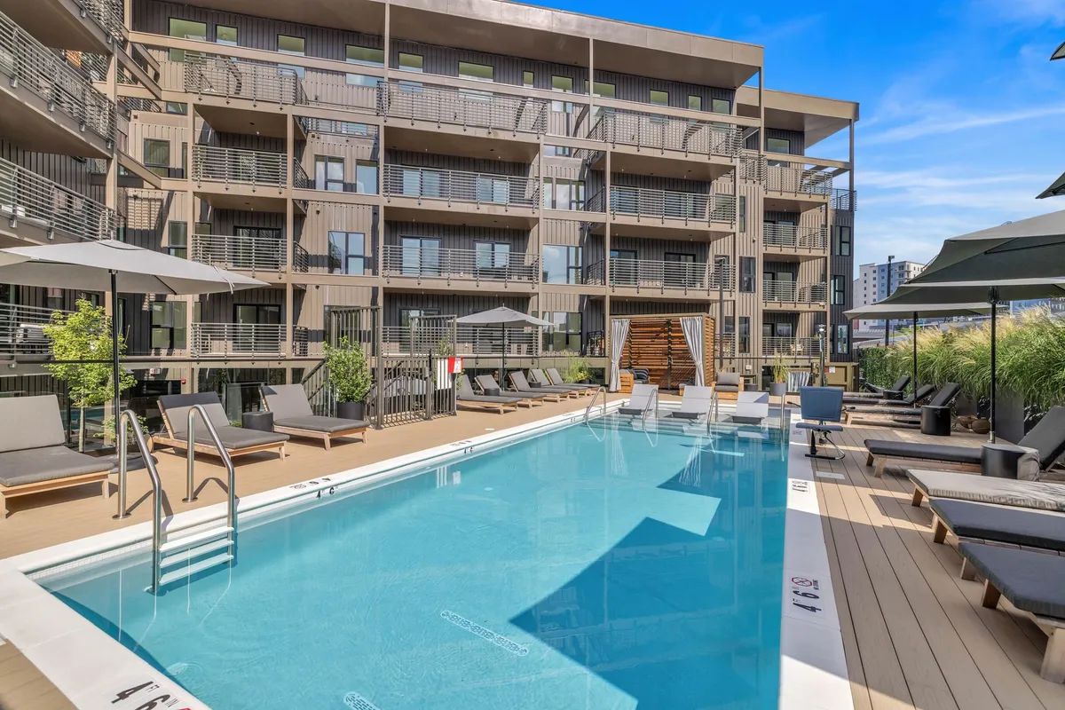 A serene outdoor swimming pool surrounded by comfortable lounge chairs and lush green landscaping, set against the building's modern facade. The scene is tranquil and inviting, perfect for sunbathing, swimming, or relaxing with friends.