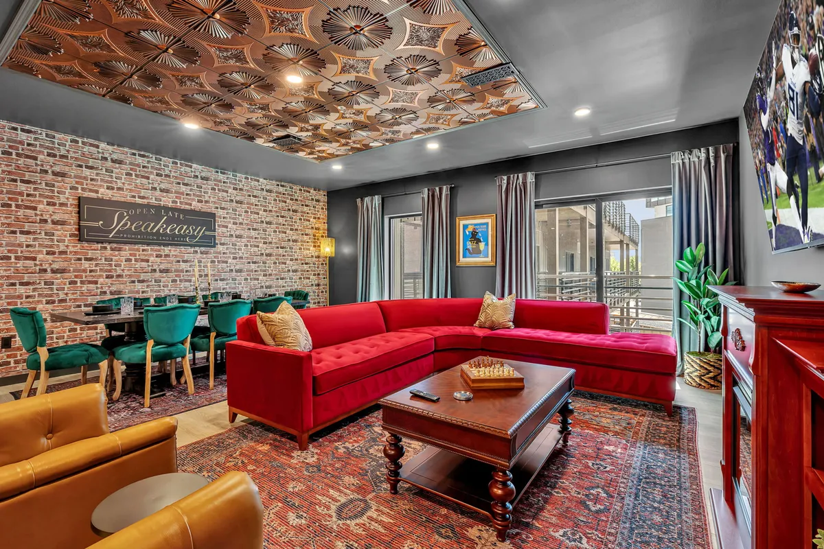 This image features a cozy living area with a bold red sectional sofa, an ornate ceiling, exposed brick wall with signage, and a tastefully arranged dining area with green chairs. The scene communicates a warm, communal environment that invites groups to relax, socialize, or enjoy a meal together.