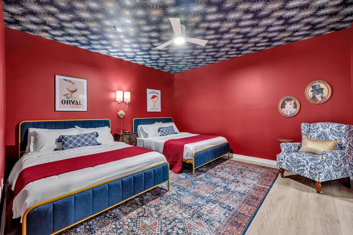 A playful yet cozy bedroom showcasing two beds with blue upholstered headboards, a vibrant red accent wall, a patterned rug, and an armchair in one corner. The space feels both fun and comfortable, ideal for family or group stays.