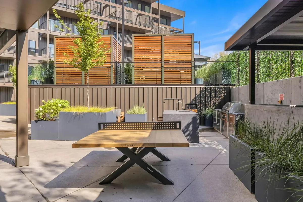 An inviting BBQ and outdoor dining area with a wooden table, surrounded by green plants and decorative screens. The space encourages social gatherings, outdoor meals, and relaxed summer evenings.
