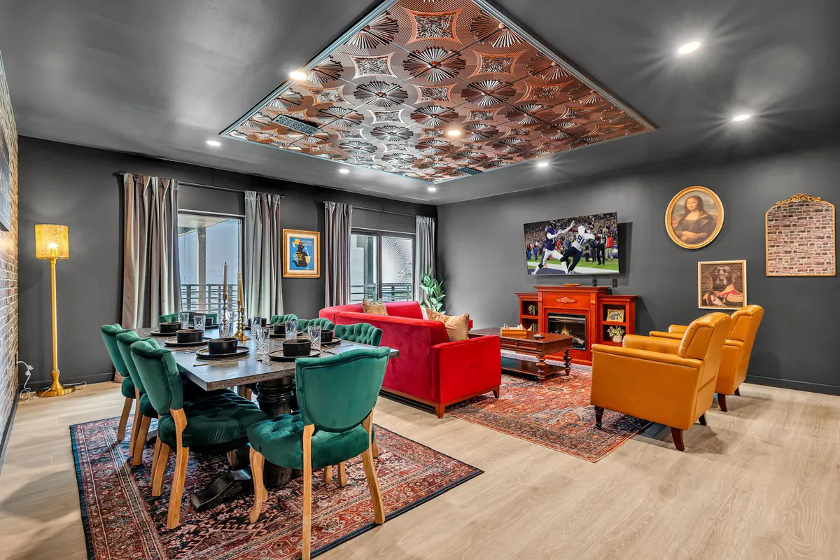The image showcases a stylish open-concept living and dining area featuring a vibrant red sofa, a sleek dining table with modern green chairs, a television displaying a sports game, and a decorative ceiling that adds vintage flair. The space is designed for gathering, celebrating, and relaxing, with plenty of room for groups to dine, socialize, or watch the big game together.