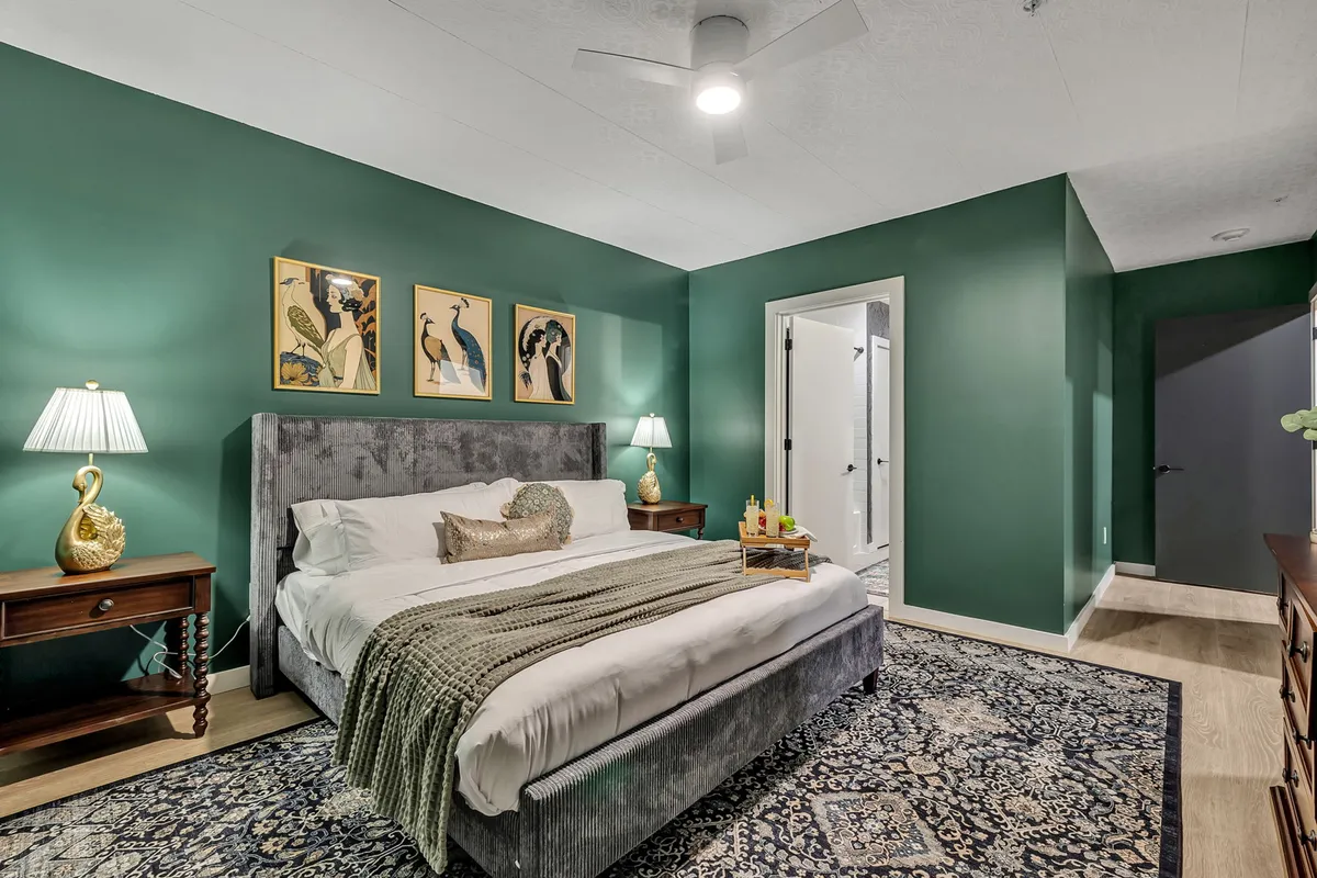 A cozy bedroom featuring a plush bed adorned with soft linens, contemporary artwork above the headboard, matching nightstands with lamps on either side, and a richly textured rug underfoot. Natural light filters in, creating an inviting atmosphere perfect for relaxation or quiet reading.