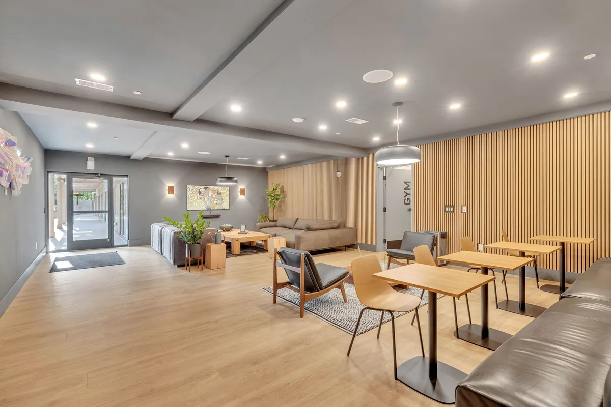 A contemporary and welcoming lounge area with comfortable sofas, wooden tables, visible gym entrance, and decorative wall art. The scene captures a communal space ideal for relaxing, socializing, or working, with a sense of professional management and communal hospitality.