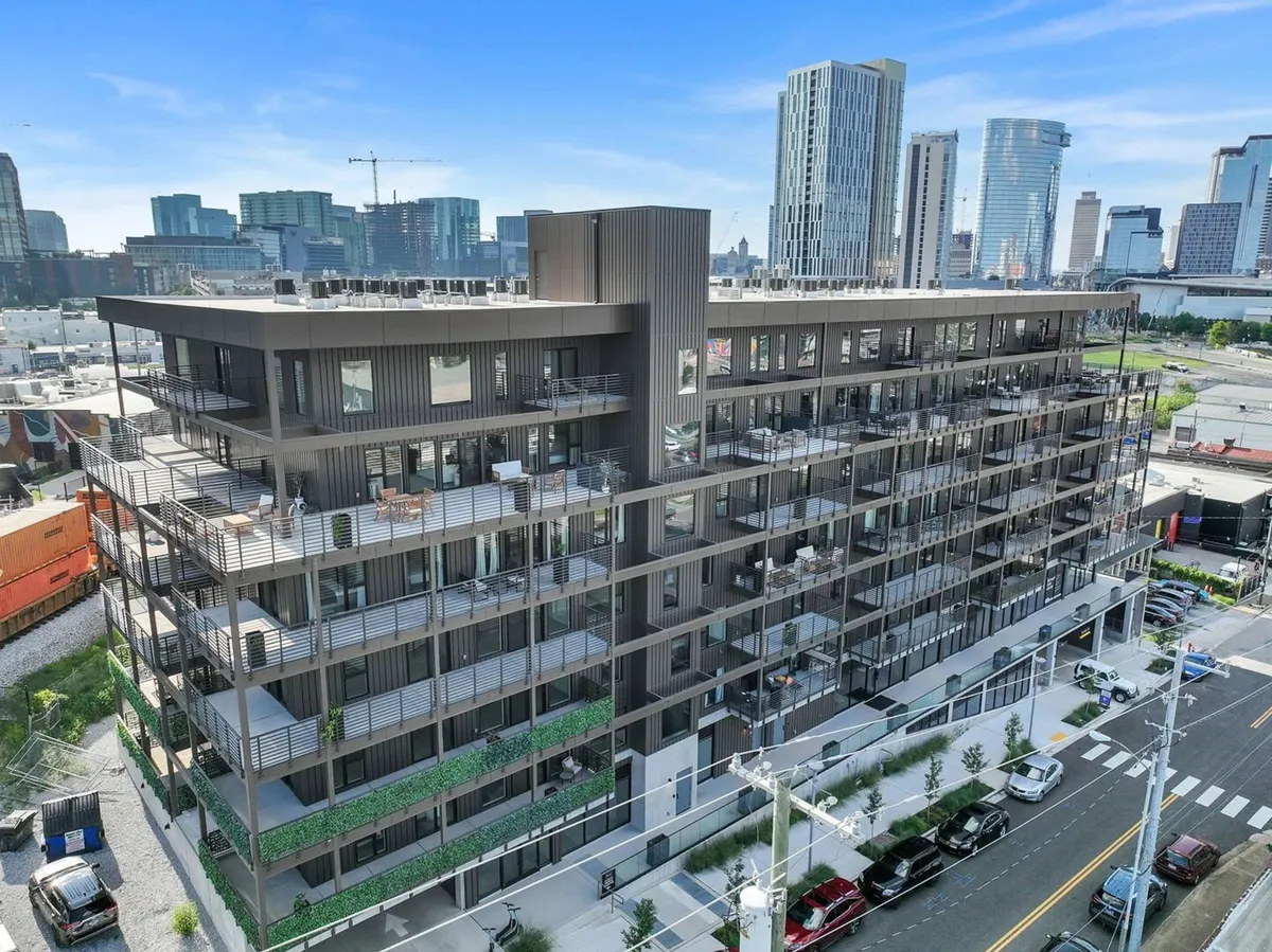 A modern apartment building with a rooftop terrace and multiple balconies furnished for outdoor gatherings. The city skyline and sunset sky in the background emphasize urban luxury and panoramic views.
