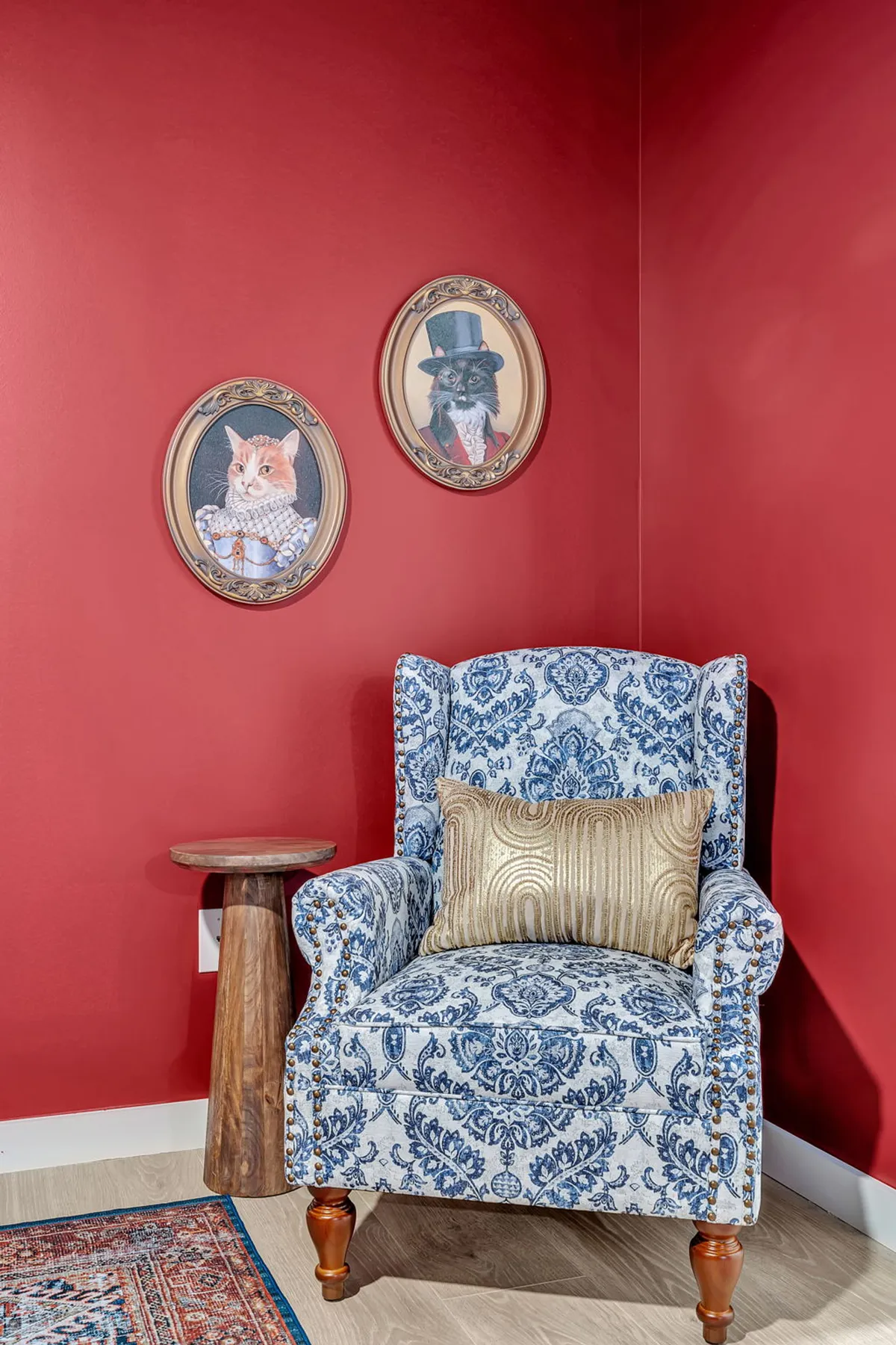 A cozy interior seating nook with a red wall, playful cat paintings, a blue patterned armchair, and a wooden side table with a gold pillow. The scene exudes a sense of comfort, relaxation, and visual charm, ideal for families and guests seeking a warm, managed environment.